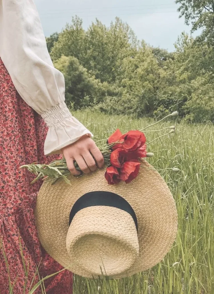 a woman in a green field holding red flowers and hat