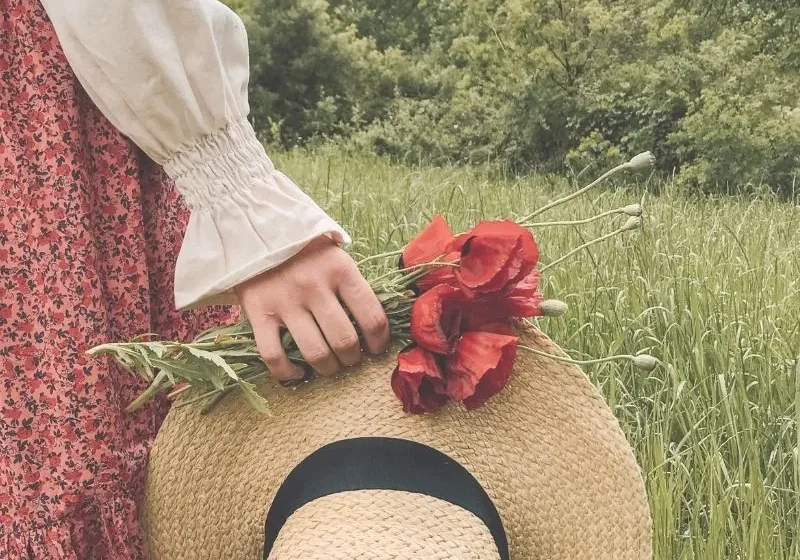 a woman in a green field holding red flowers and hat