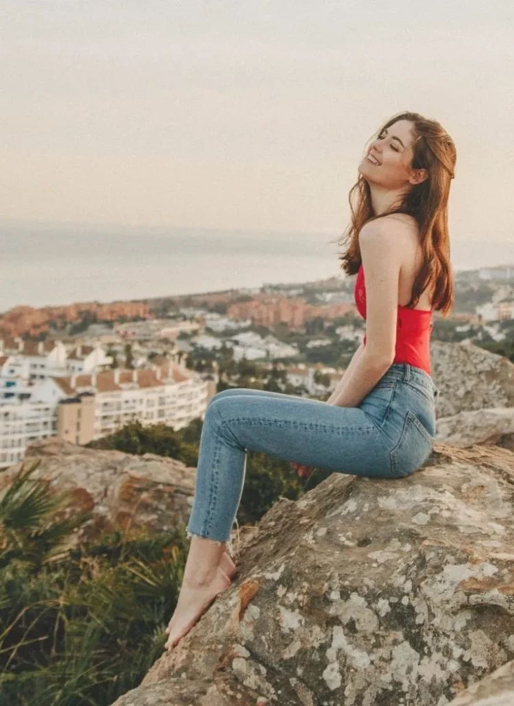Woman in red top and denim pants sitting on rocks with view
