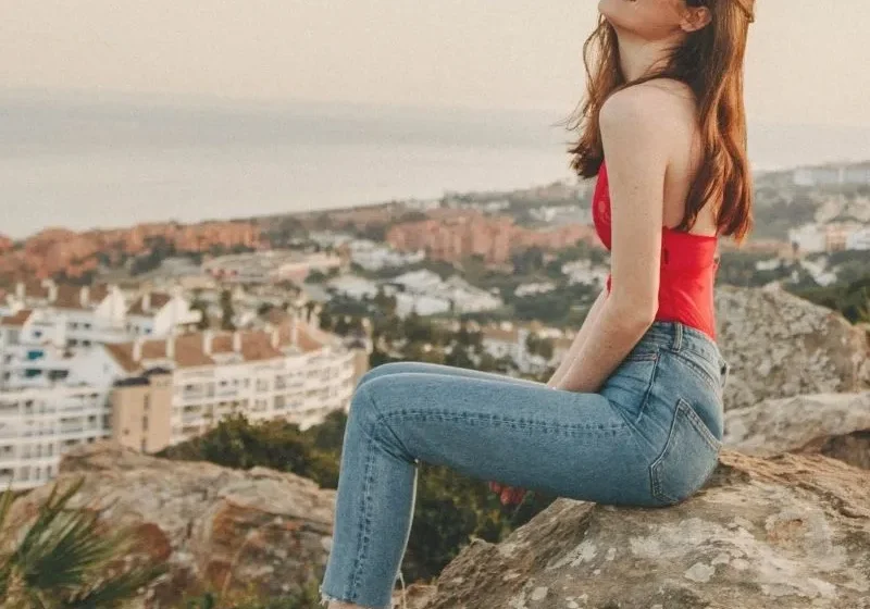 Woman in red top and denim pants sitting on rocks with view