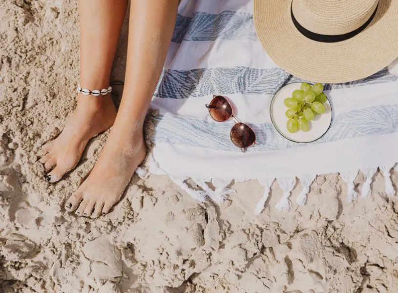 a woman's feet on beach towel with sunglasses, grapes and hat