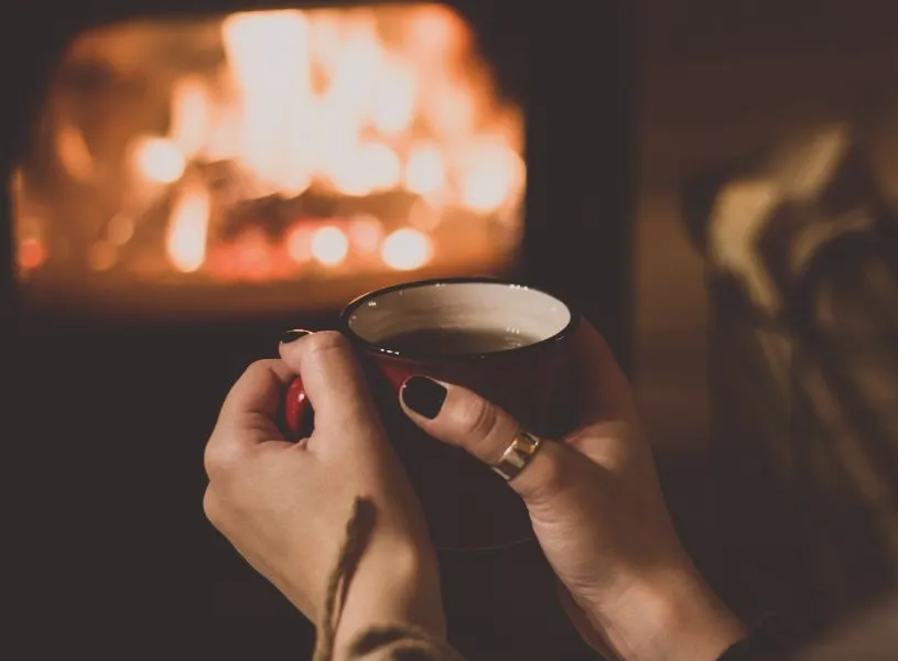 woman holding hot tea next to fireplace - a winter bucket list activity to do for adults