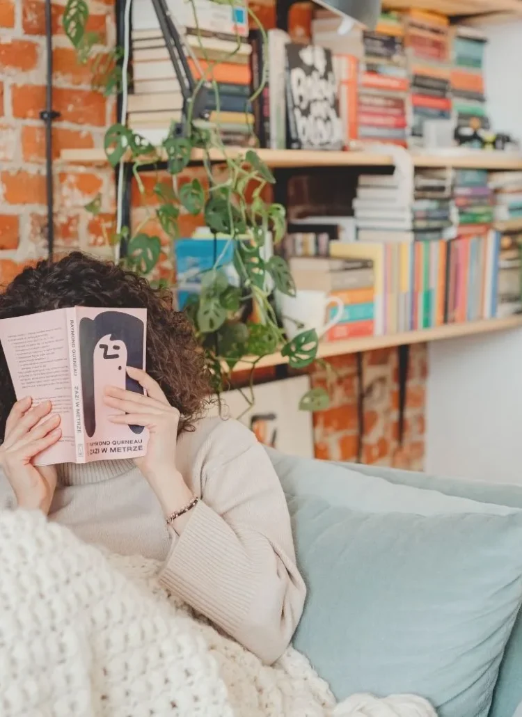young woman sitting on the couch hiding her face behind a pink book