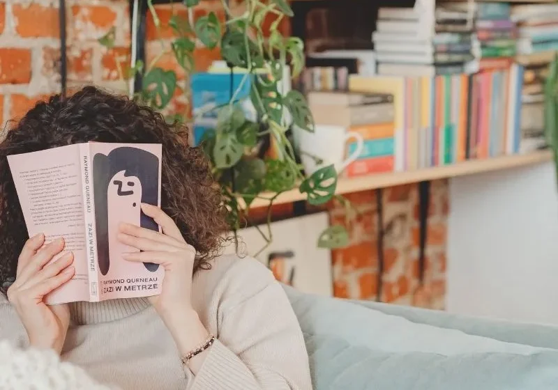 young woman sitting on the couch hiding her face behind a pink book