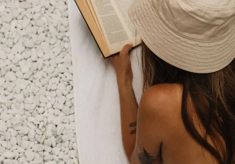 woman enjoying her summer solo moments. She's laying down in bathing suit and hat reading a book