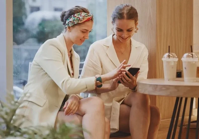 two ladies looking at the phone on a Friday smiling and chatting with coffee