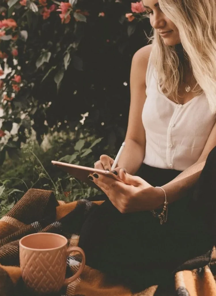 woman sitting on top of a blanket with flower bush behind her holding a tablet and pen. There's also a cup on the blanket