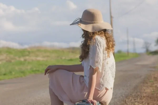woman with big hat sitting on a suitcase next to the road