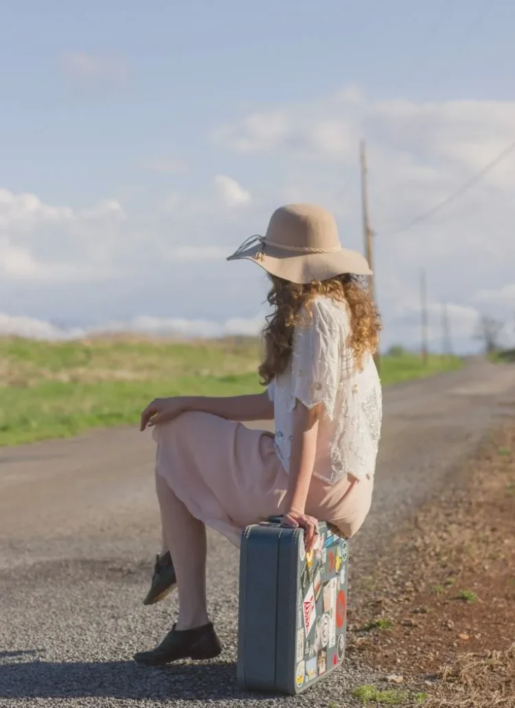 woman with big hat sitting on a suitcase next to the road