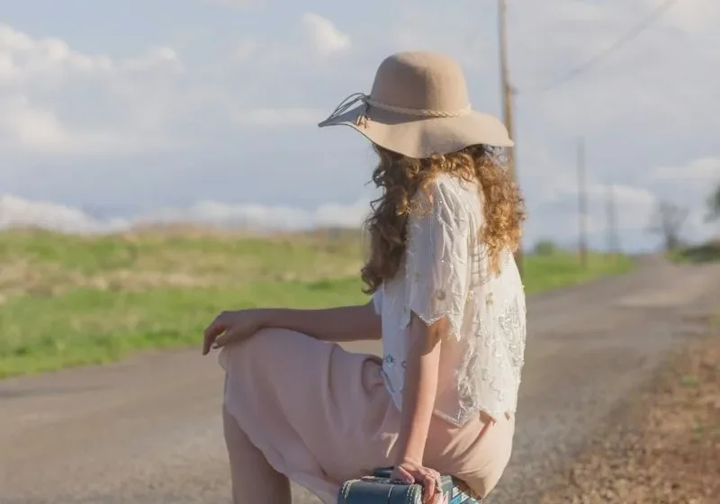 woman with big hat sitting on a suitcase next to the road