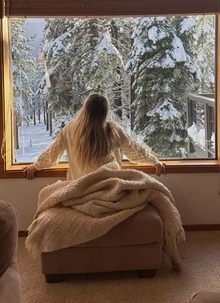 photo of woman in her 30s looking out the window with trees covered in snow